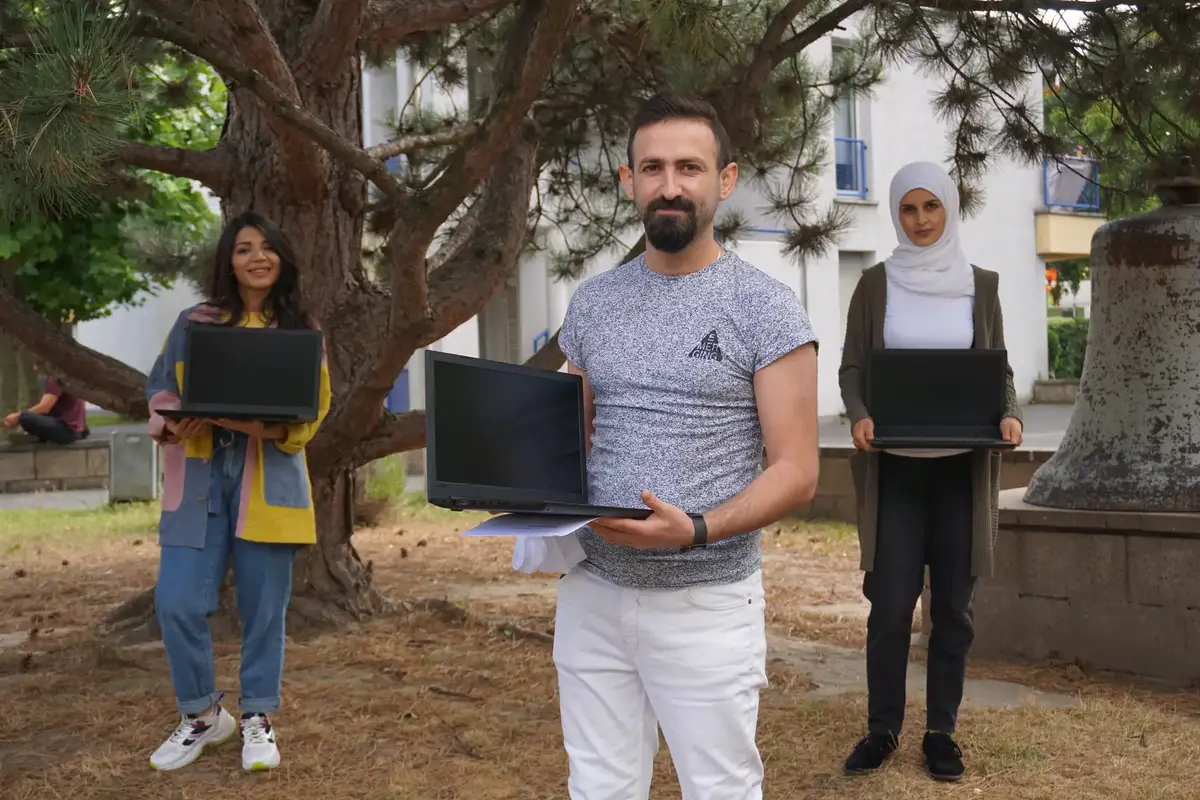 Ranim Kordy, Shear Fares und Hadiya Hussein stehen vor einem Gebäude und halten jeweils einen Laptop in den Händen