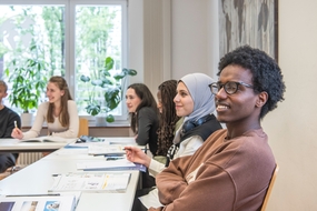 Gruppe von GF-H Sprachkursteilnehmenden im Klassenzimmer im Deutschunterricht. Die Teilnehmenden sitzen in an Tischen, lächeln und haben Unterrichtsmaterial vor sich liegen. 